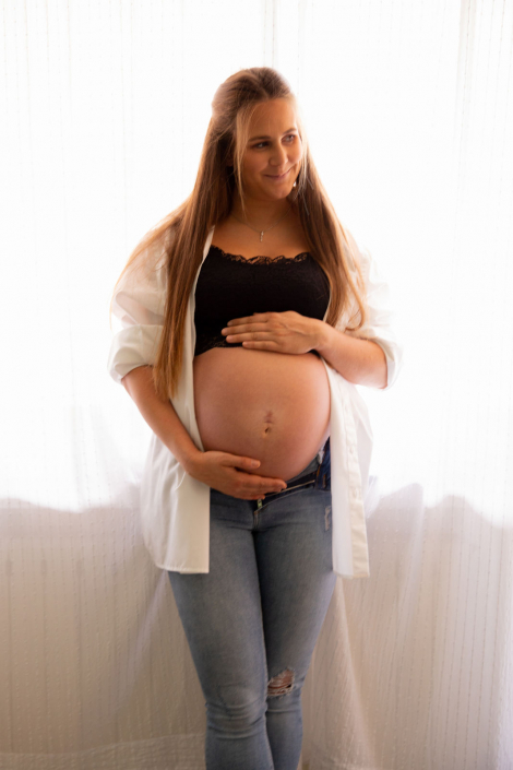 Eine schwangere Frau steht mit offenem Hemd in einem hellen Raum. Sie ist glücklich und lächelt.