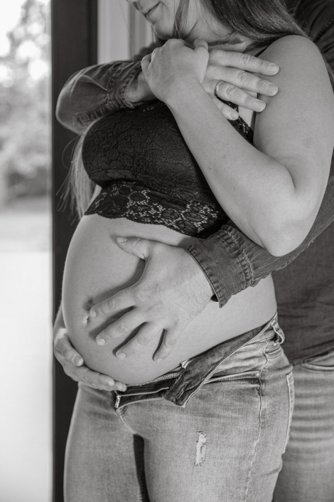 Eine schwangere Frau steht mit Ihrem Mann in einem Raum. Der werdende Vater hält die Hände stolz auf den Bauch.
