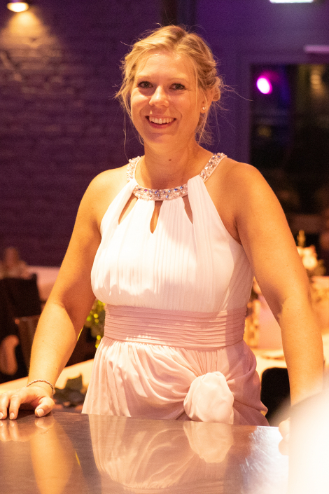 Eine glückliche junge Frau in einem Kleid steht an einem Tisch. Sie lächelt in die Kamera.