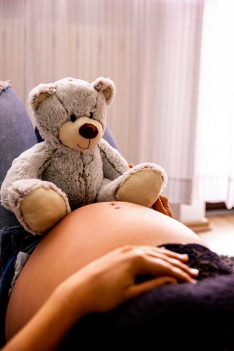 Eine schwangere Frauliegt mit offenem Hemd in einem hellen Raum. Sie ist glücklich und lächelt. Ein Kuscheltier Bär liegt auf dem nackten Bauch.