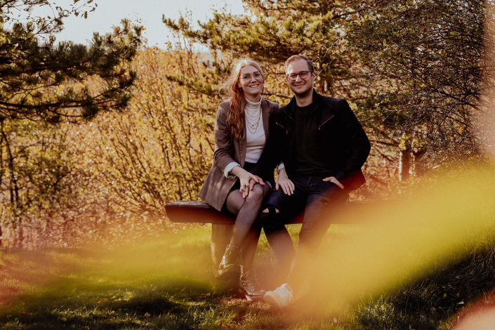 Ein junges Paar sitzt auf einer Bank bei einem Fotoshooting. Beide lächeln und haben die Hände gegenseitig verschlungen.